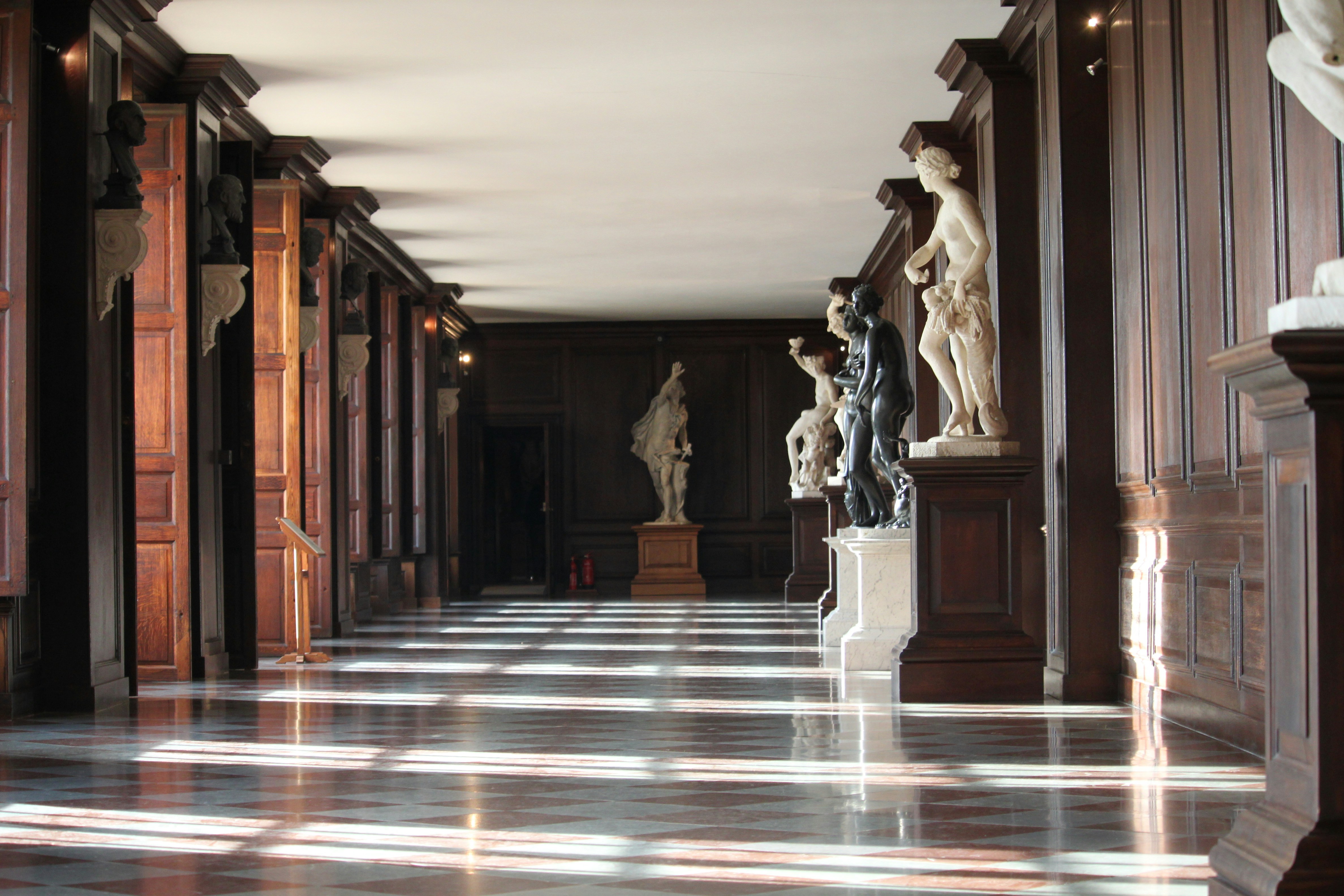 Statues line one of the long window-lit halls of Hampton Court Palace in London.