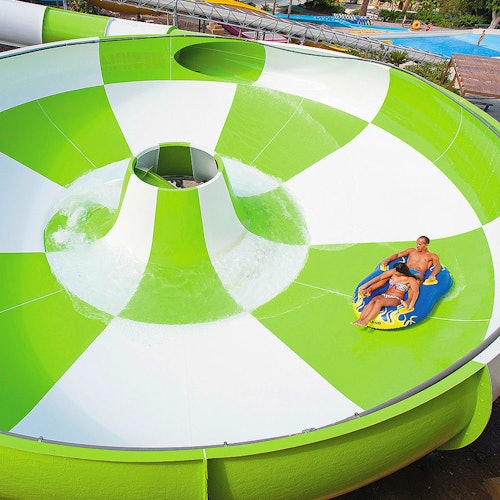 People riding an inflatable raft in a large, green and white water slide bowl at an outdoor water park.