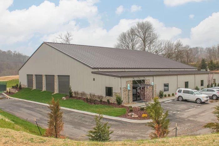 A modern, single-story building surrounded by grass and trees, with a parking area in front under a cloudy sky.