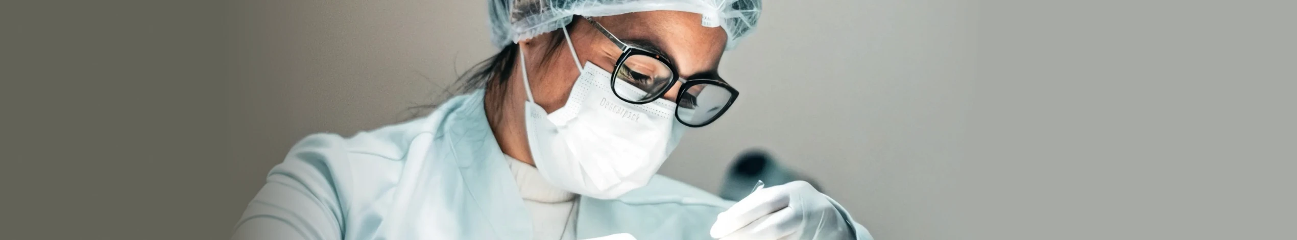 Dentista Nathália Dias em clínica odontológica usando máscara, touca e luvas durante procedimento dental, transmitindo segurança, higiene e atendimento profissional.