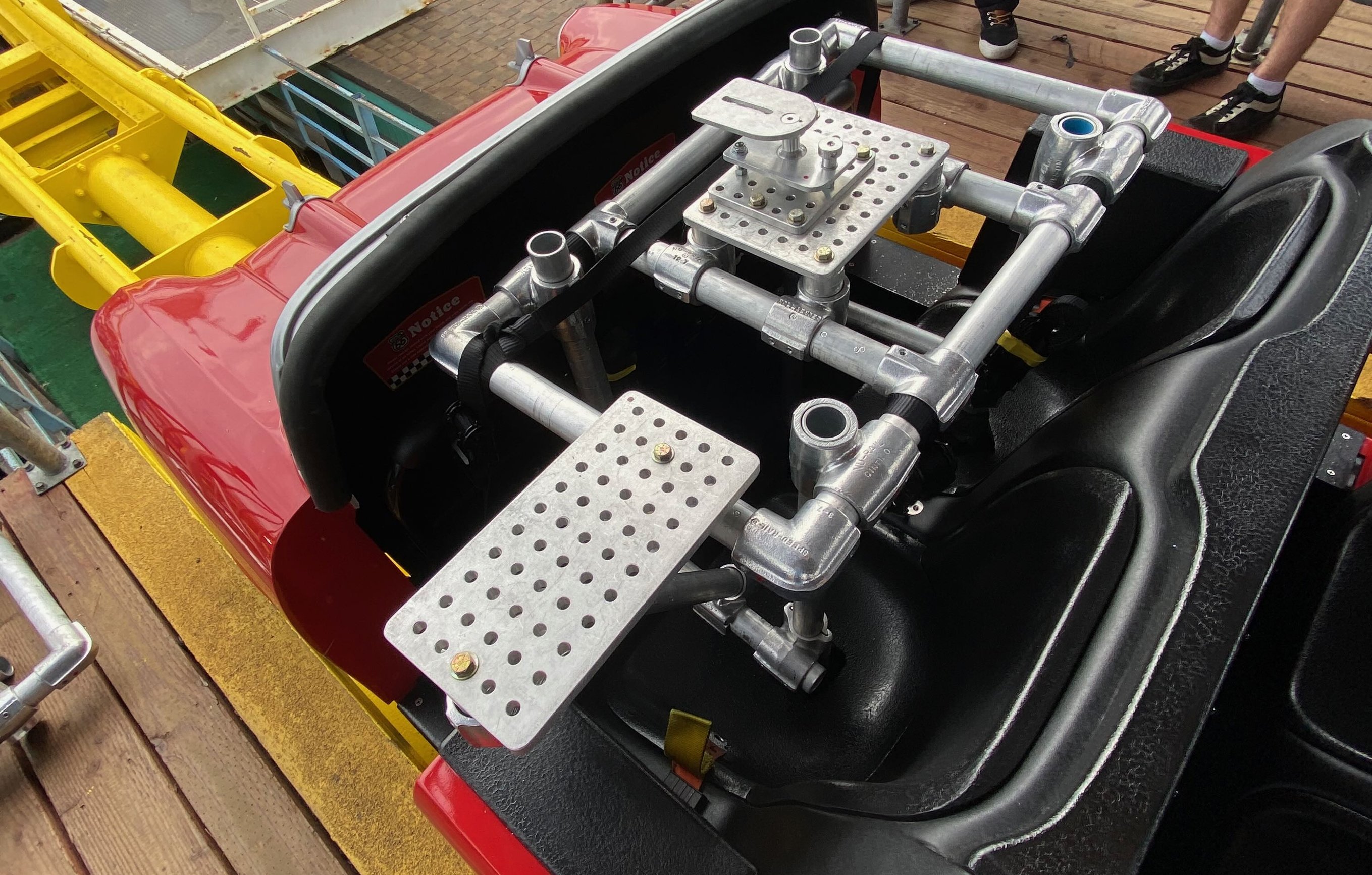 Bare speedrail and Hollaender frame with two cheeseplates built into the seat area of a red Santa Monica Pier roller coaster car, with the yellow coaster track structure and pier boardwalk visible in the background