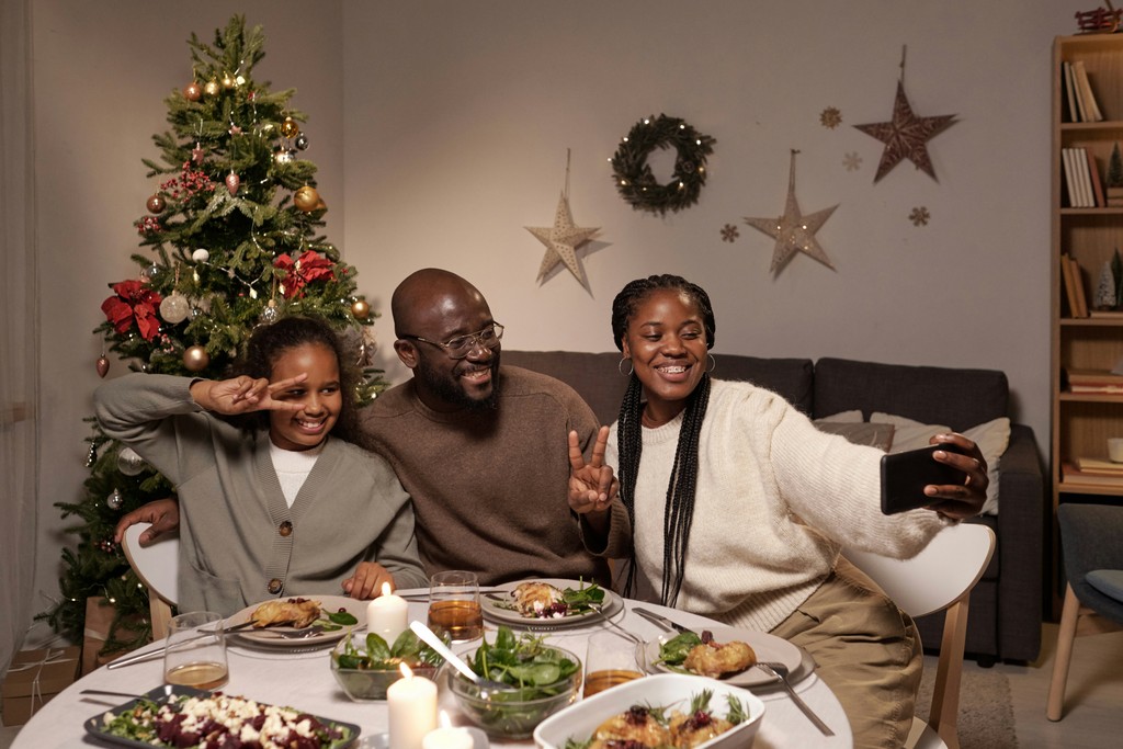 A joyful family takes a selfie during Christmas dinner, featuring a decorated tree with ornaments and lights in the background, evoking the festive holiday spirit.