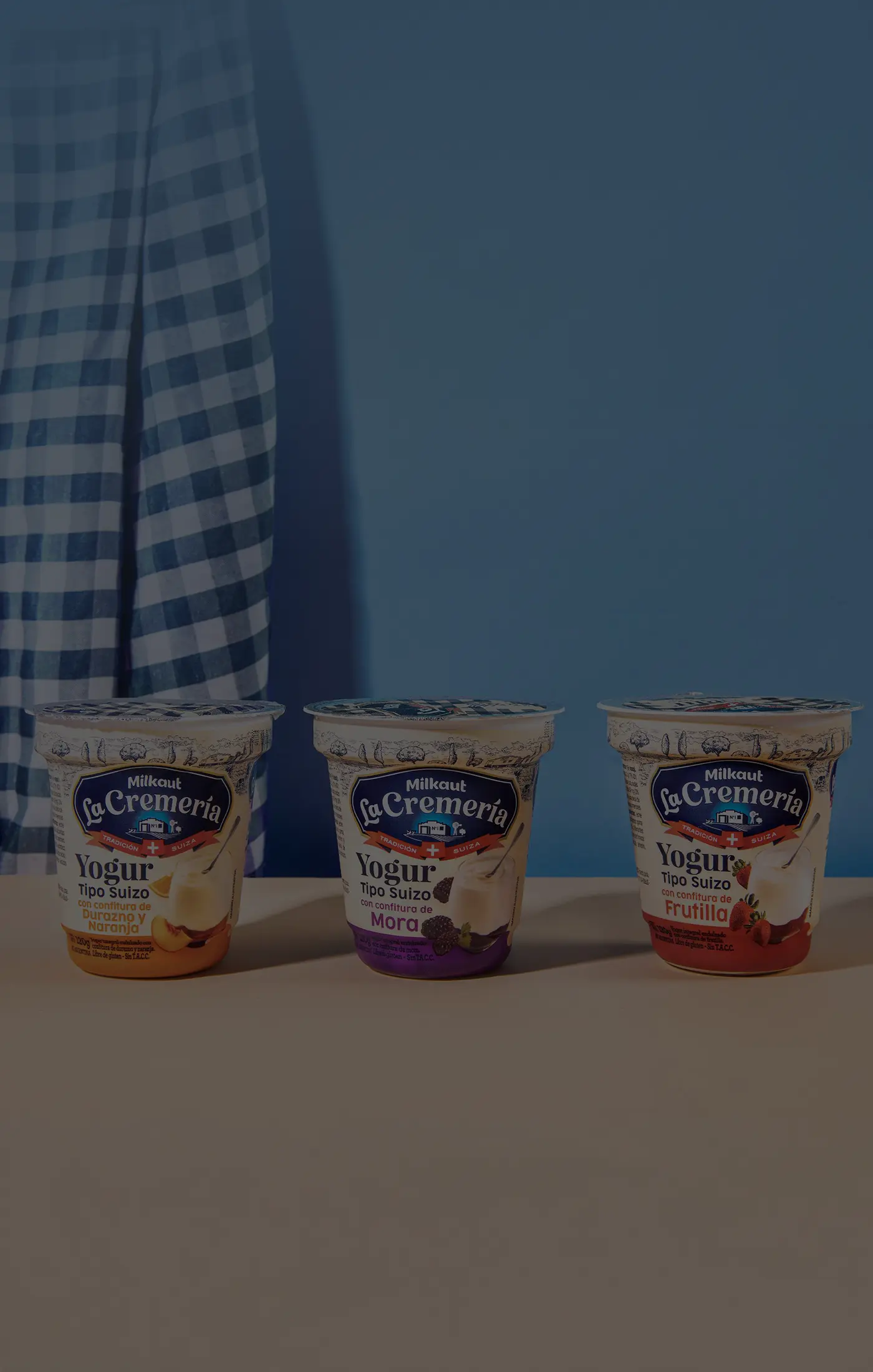 Three yogurt containers of different flavors—banana, blackberry, and strawberry—are arranged on a beige surface. A blue gingham cloth hangs in the background.
