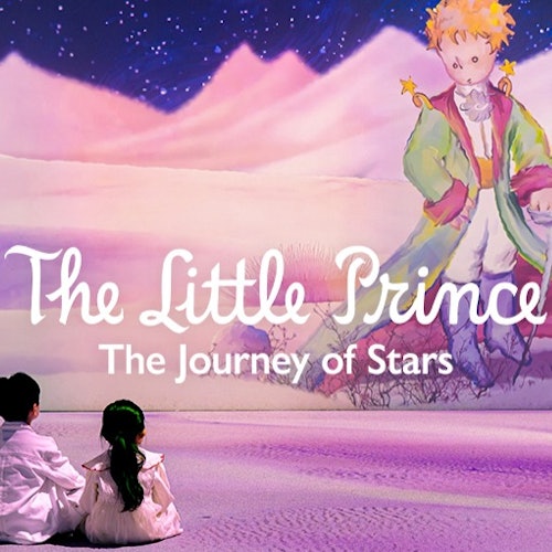 两个孩子坐在一个色彩缤纷的展览前，展览文字为“小王子：星星之旅”，并展示了这个角色的大型壁画。