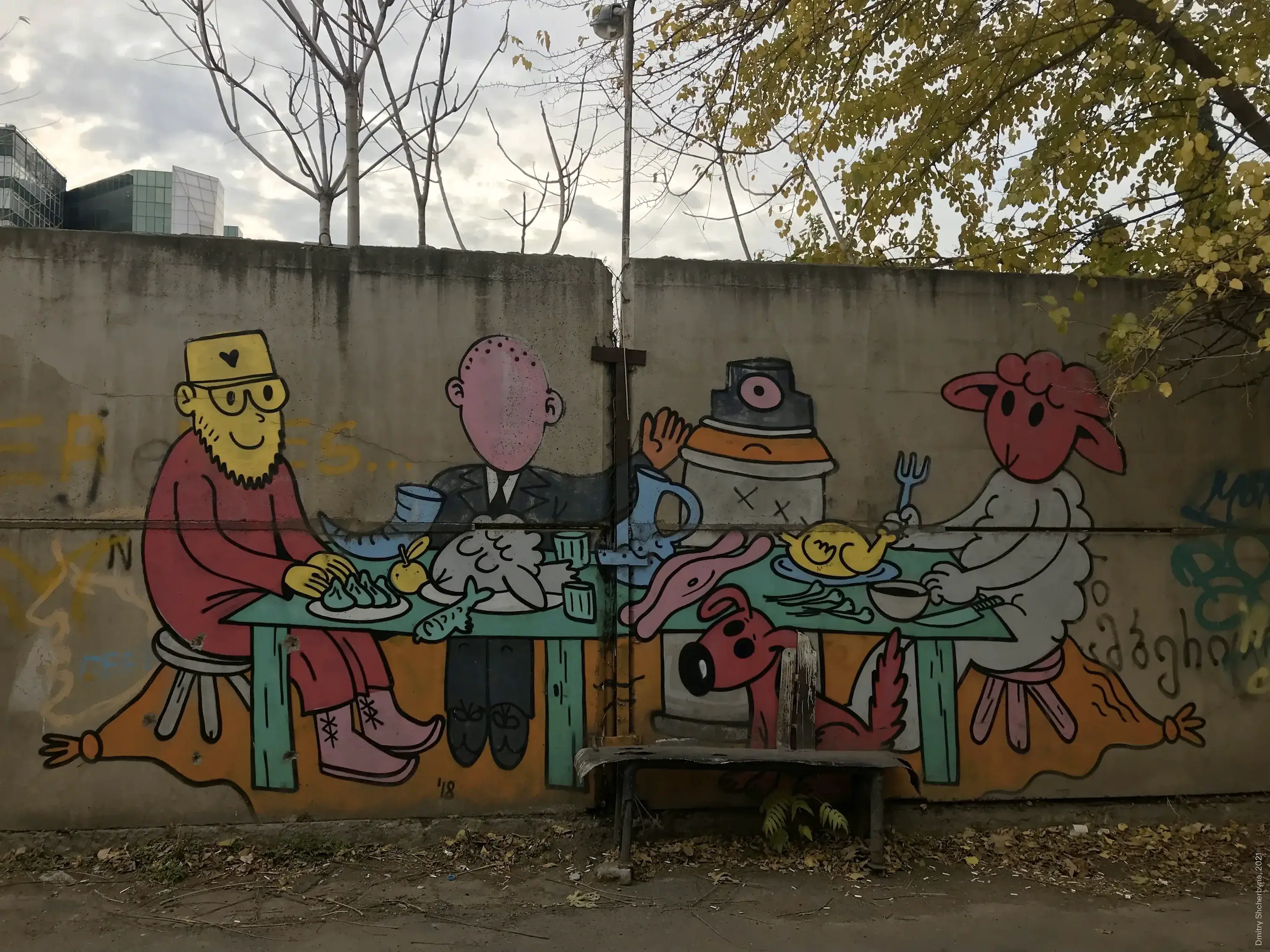 A colorful cartoon mural on a concrete wall shows four quirky characters sitting around a table laden with food and drinks — a bearded man in a red robe and yellow hat, a bald man in a suit, one-eyed robot wearing a top hat and a pink sheep holding a fork. A red dog peers out from under the table.