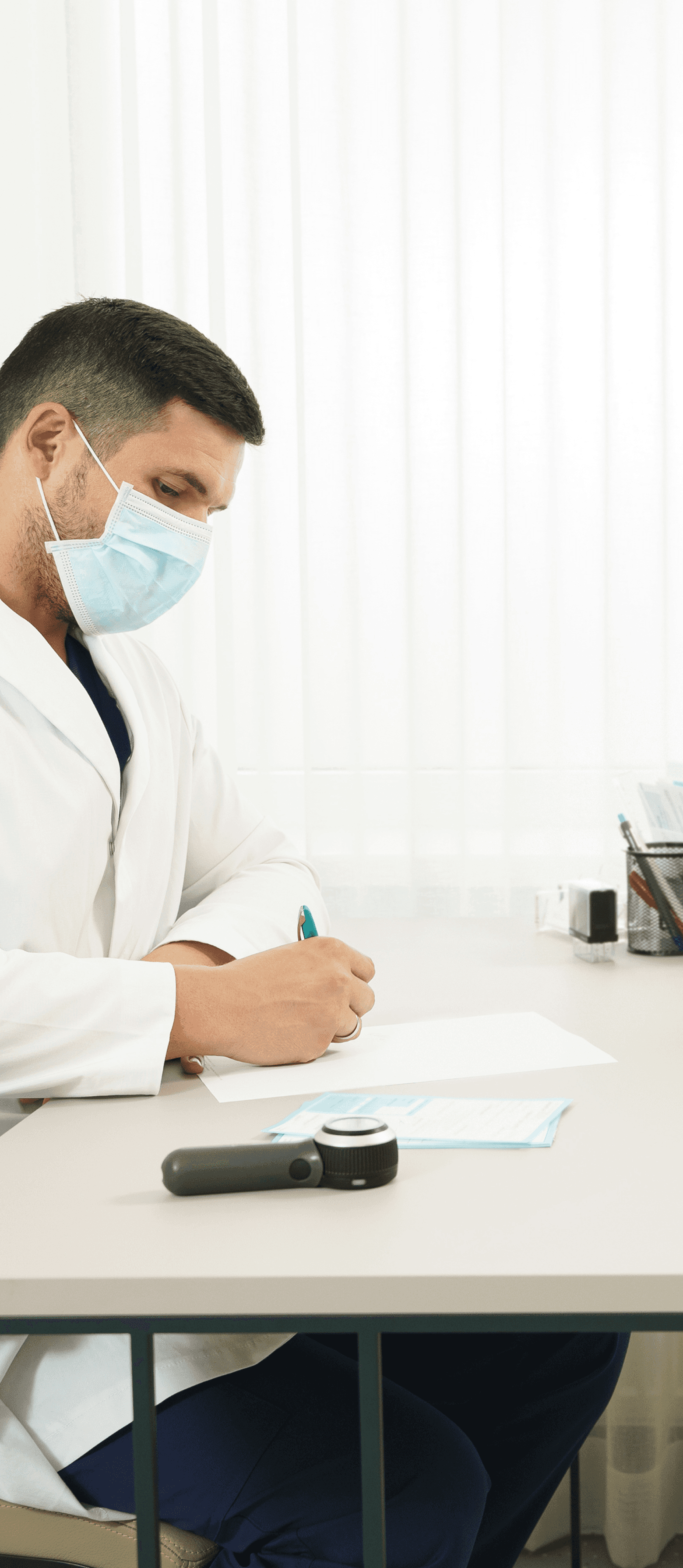 A doctor in a white coat, wearing a mask, writes notes at a desk while a patient in a striped shirt, also masked, attentively listens in a well-lit room.