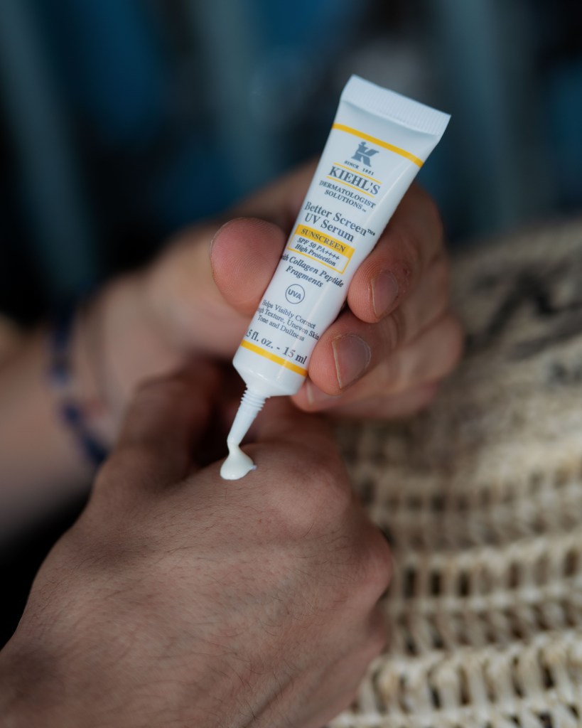 A person applying Kiehl's Better Screen UV Serum sunscreen to their hand, with the product tube prominently displayed.