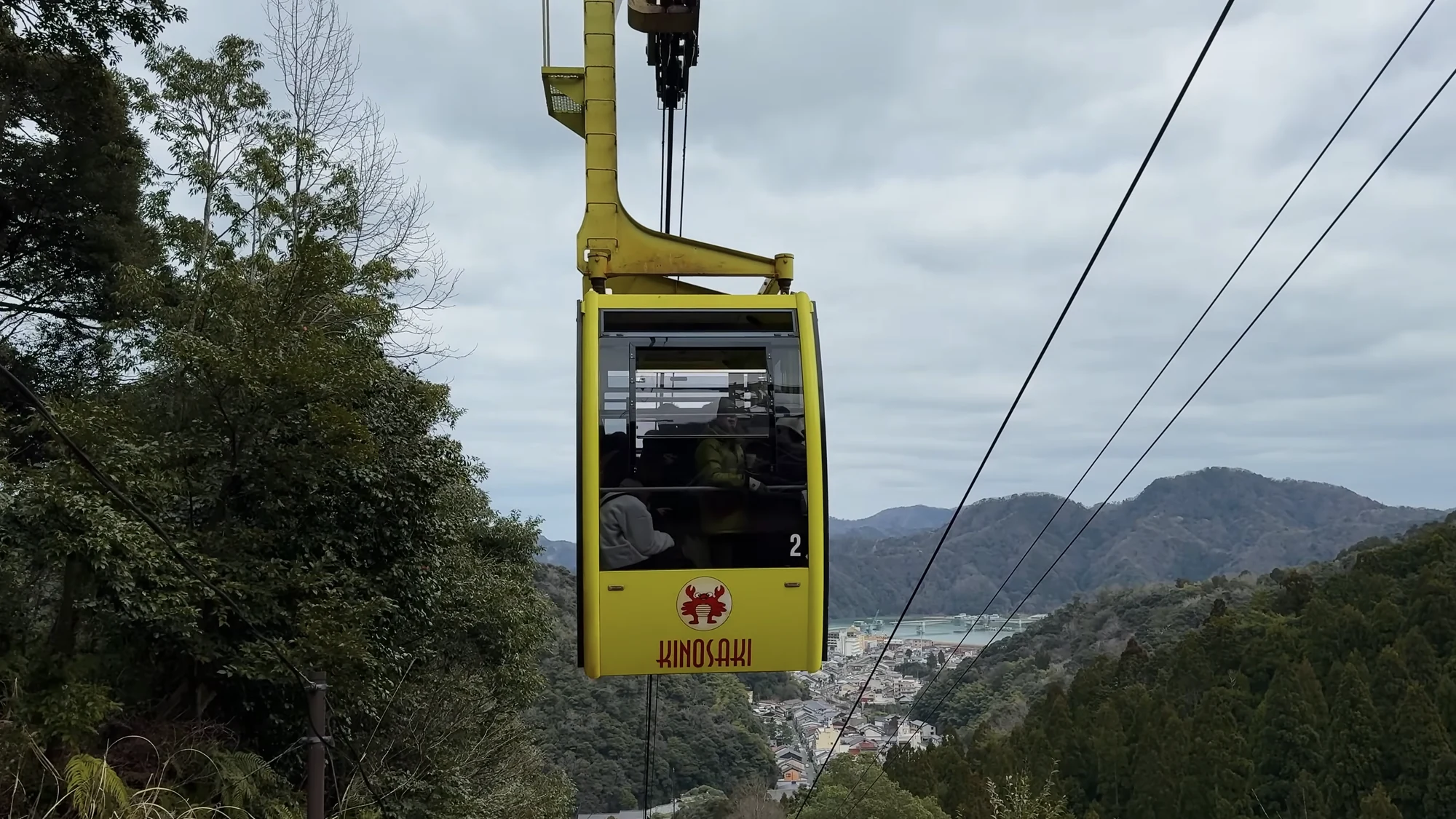 A yellow ropeway gondola climbing above wooded hillside with the town below.