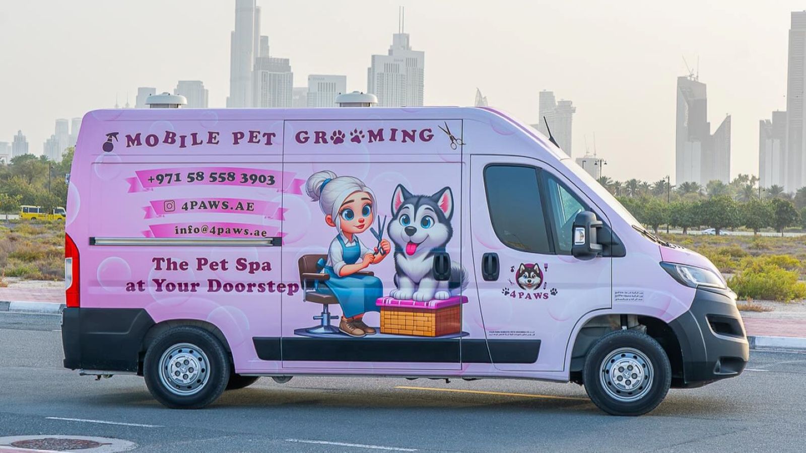 A pink mobile pet grooming van in the middle of the road with skyscrapers in the background.