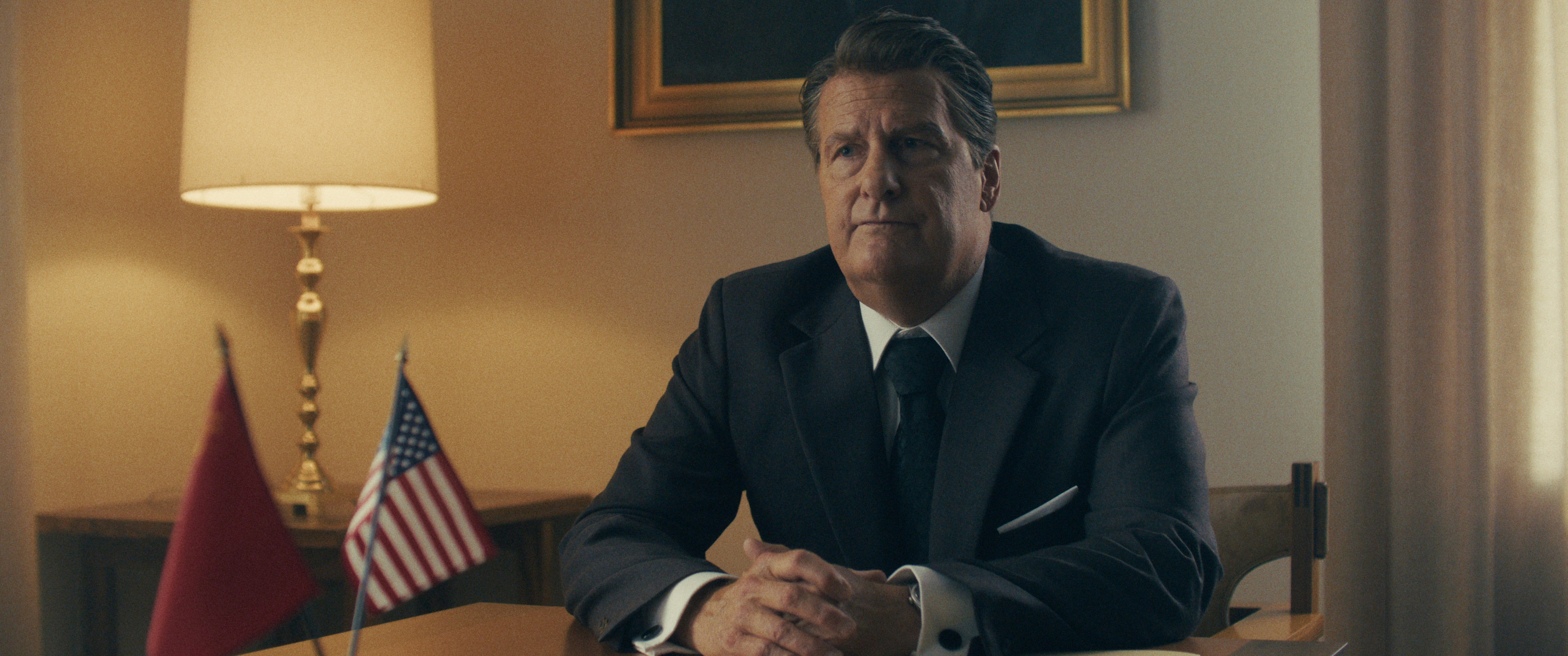 Actor Jeff Daniels as Ronald Reagan sits at a table with his hands folded, a brass lamp and gilt-framed painting behind him, and small Soviet and American flags in the foreground.  Photo: Courtesy of Angel Studios
