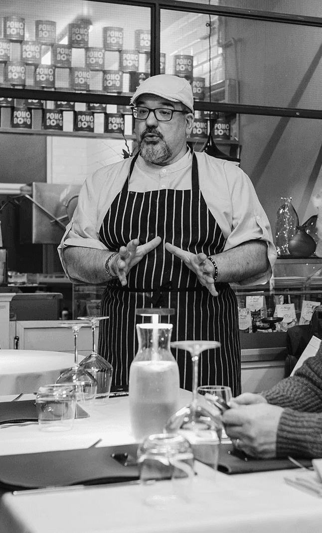 A chef wearing a striped apron, light-coloured shirt and cap speaks to diners while gesturing with his hands. He stands in a restaurant setting with glassware and a water carafe on the table in front of him, and shelves of ingredients and packaging visible in the background.