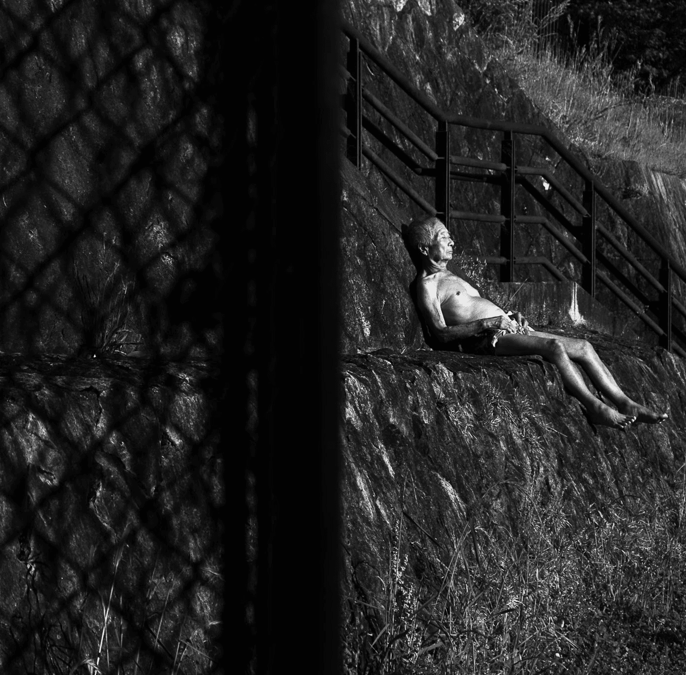 An older man with a bare chest reclines on a sloping stone embankment in the sun while the shadow of a chain-link fence fills the foreground.