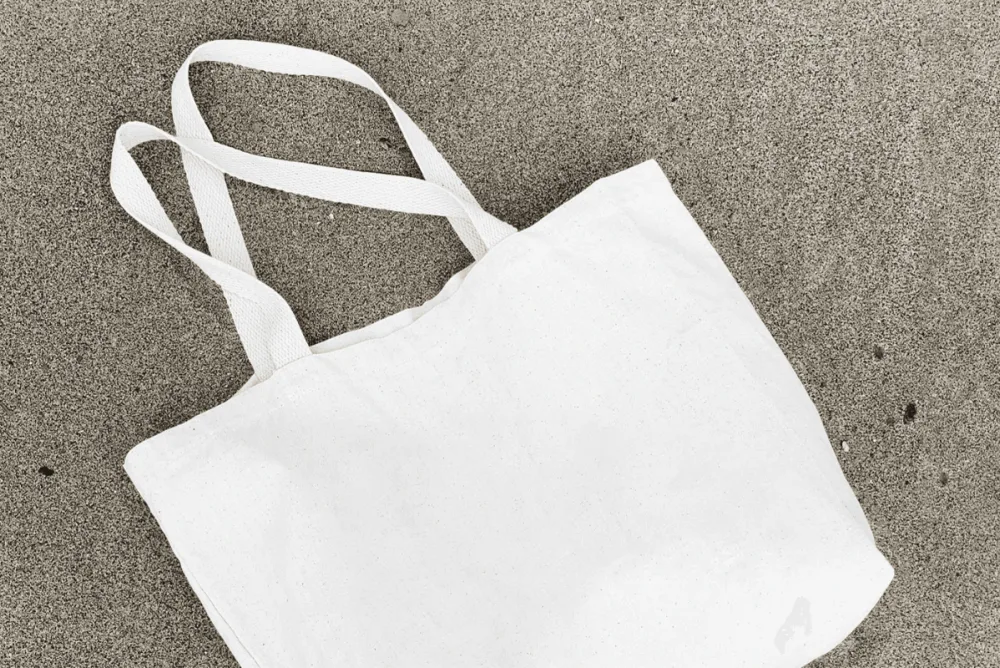 White cotton tote bag mockup lying on a sandy surface. The bag features sturdy fabric handles and a blank canvas ideal for showcasing custom designs, logos, or branding.