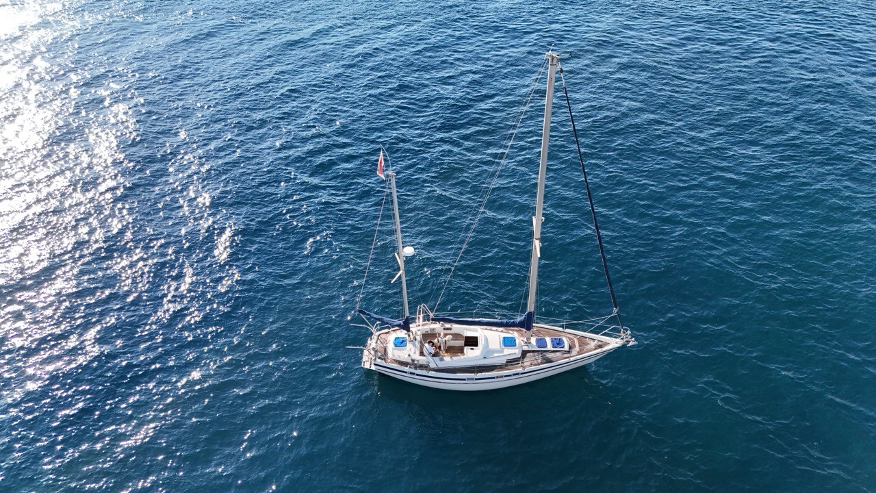 Aerial drone top-down view of a Contest 38 Ketch sailboat at anchor on sparkling deep-blue Mediterranean sea — white-hulled offshore cruising ketch with blue sail covers and Maltese flag, sunlight reflecting off open water