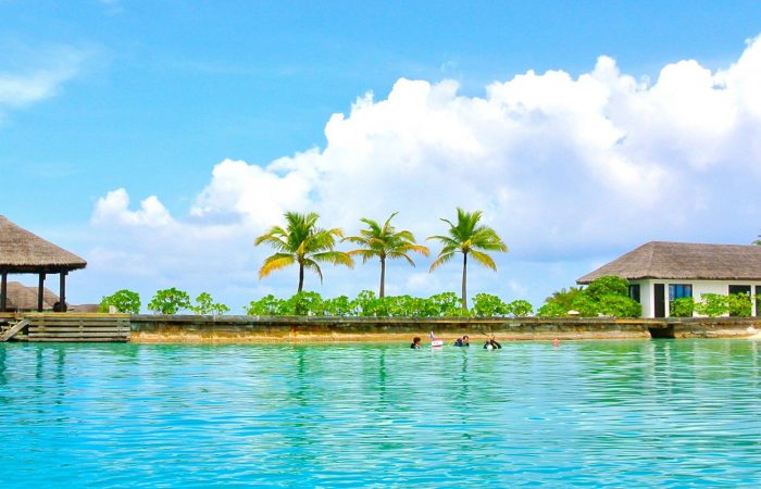 A rustic outdoor dining table set with wine glasses, cutlery, bread, and olives sits amidst lush vineyard rows, with a historic Tuscan villa in the background under a serene sky, capturing the essence of a picturesque Italian countryside landscape.