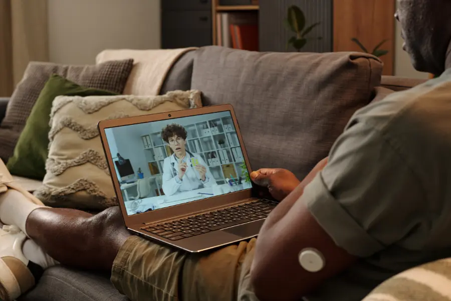 Man on a sofa using AI medical assistant software to consult a doctor via laptop video call from home.
