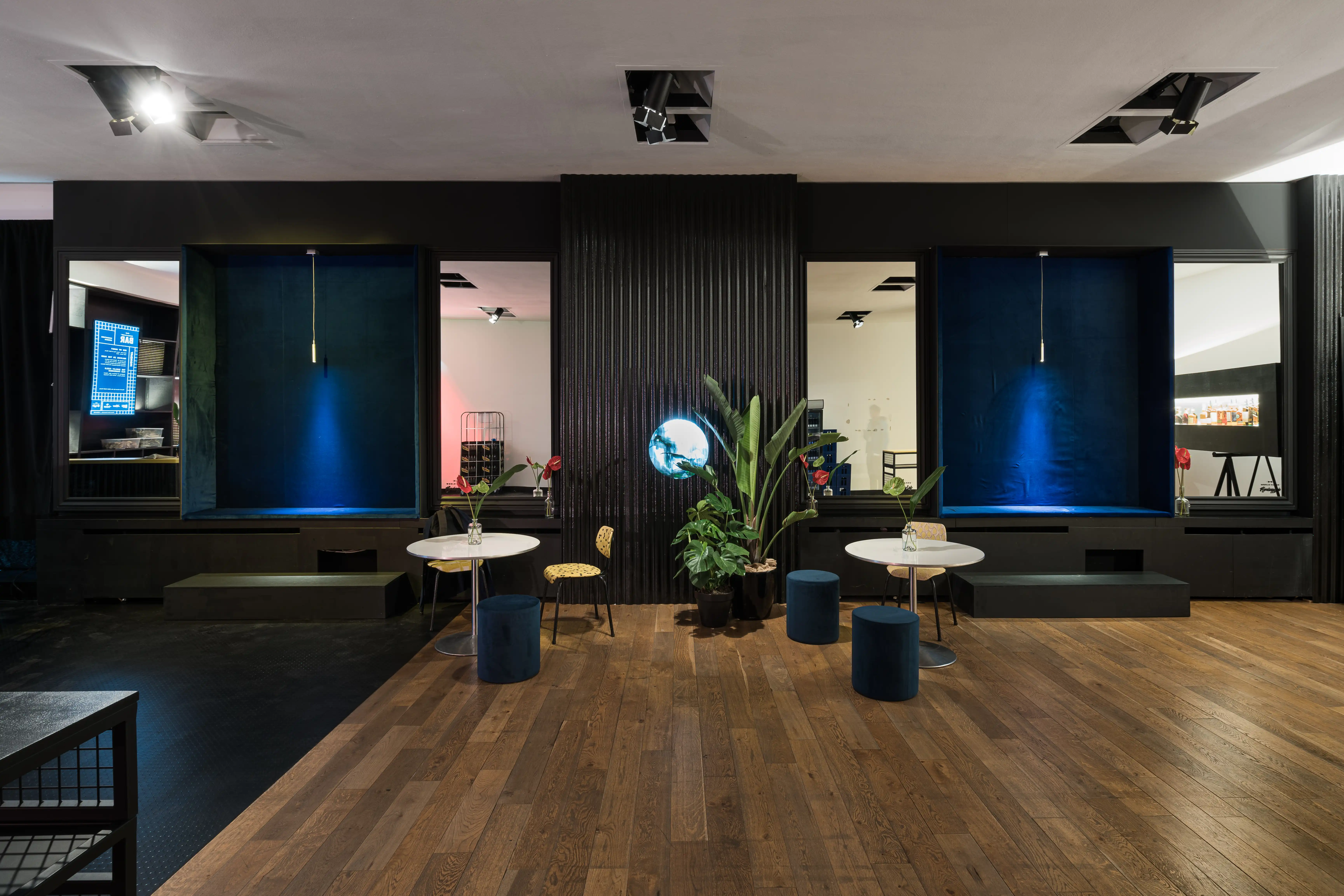 Lounge at DIAGEO BCB19 with black walls, two blue-velvet display niches, marble bistro tables, navy poufs and plants on wooden flooring.