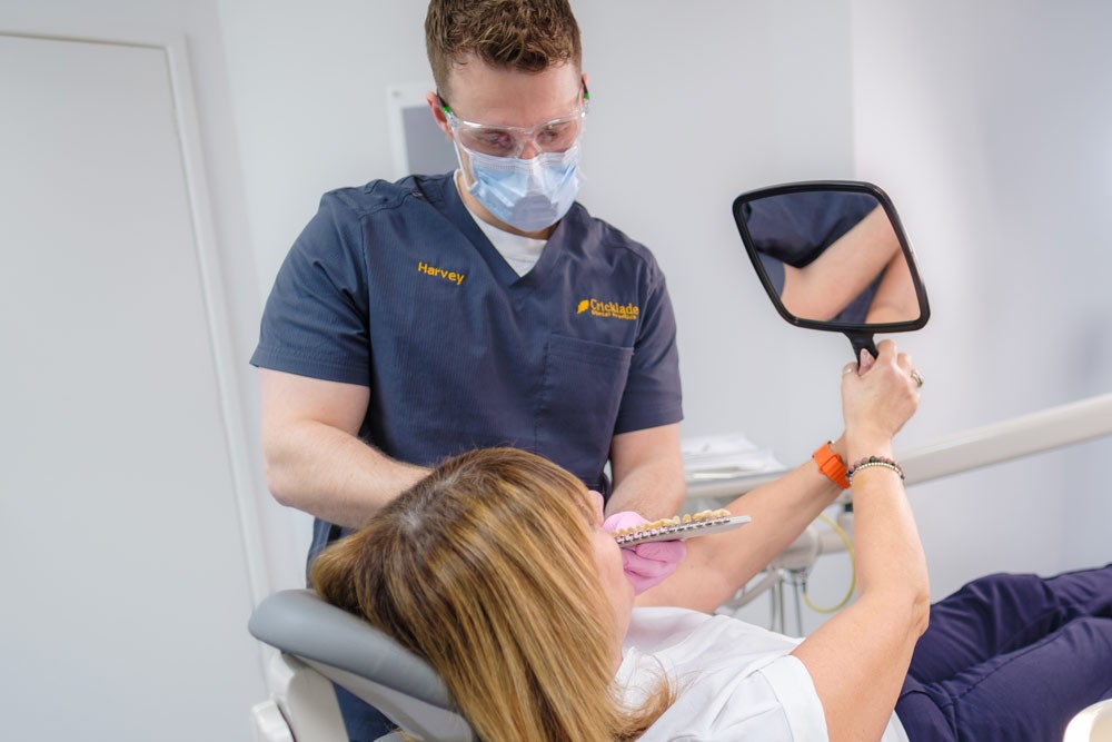 A dentist wearing protective eyewear and a mask consults with a patient holding a mirror while matching a dental shade guide to her teeth in a modern dental clinic.