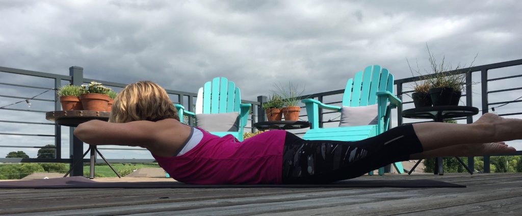 Heather Hausman is practicing yoga outdoors on a wooden deck, performing a Superman pose with overcast skies in the background, surrounded by blue chairs and potted plants.