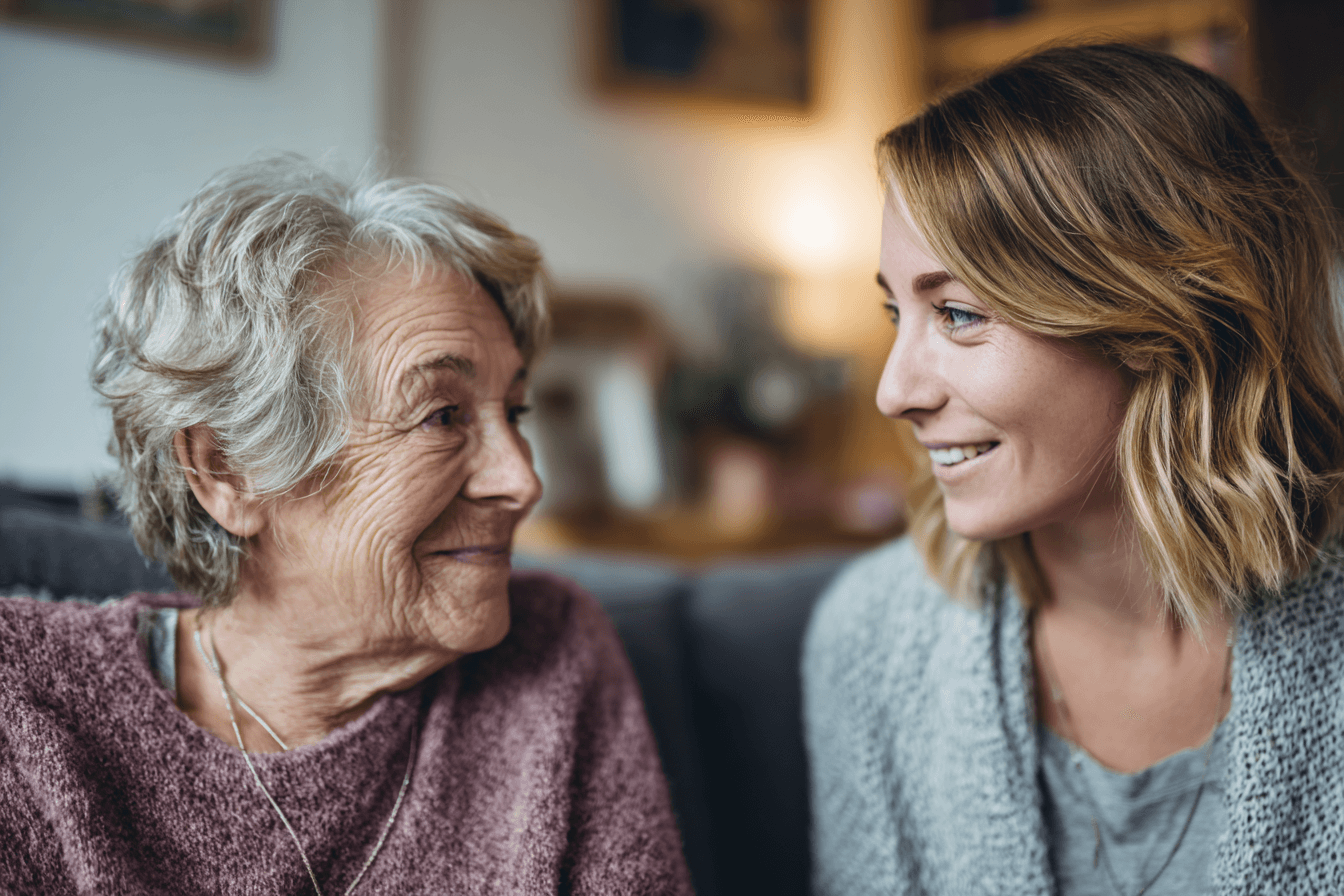 a home care agency marketer (female, 40s), talking with an adult female in her elderly mother's home. They are sitting down and smiling.