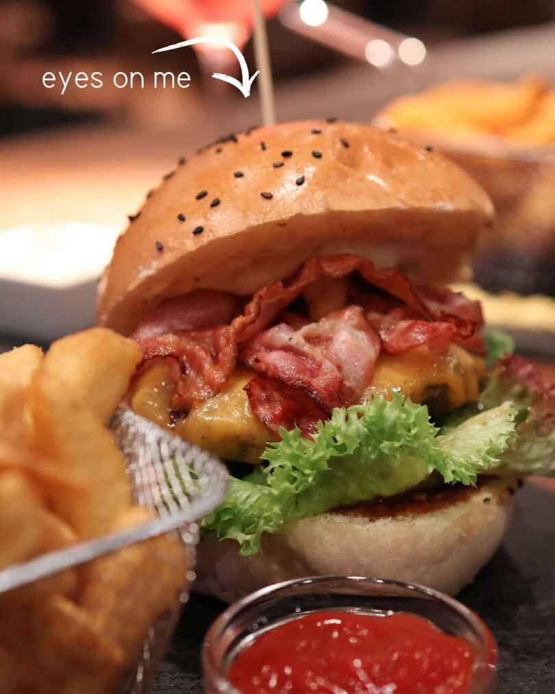 A close-up photo of a bacon cheeseburger with a sesame bun, melted cheese, crispy bacon, and lettuce, served with fries and ketchup on the side.