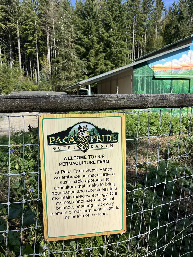 Alt text: Sign on a fence at Paca Pride Guest Ranch welcoming visitors to a permaculture farm, with forest and farm buildings in the background.