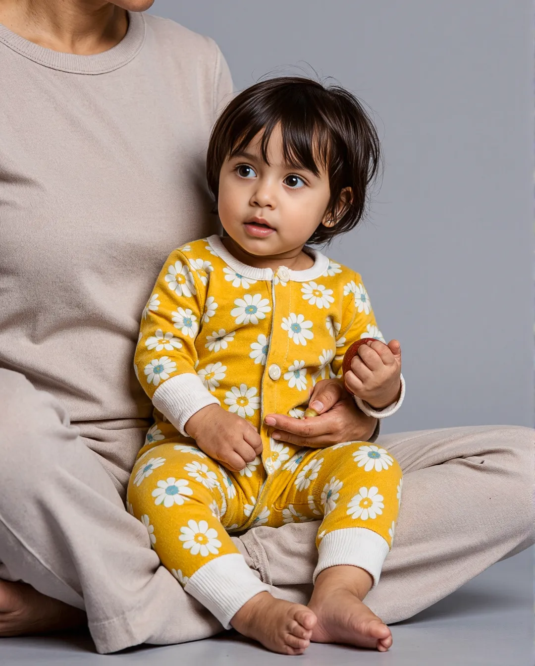 Child wearing bright yellow daisy-patterned outfit sitting with adult in neutral clothing against light gray background
