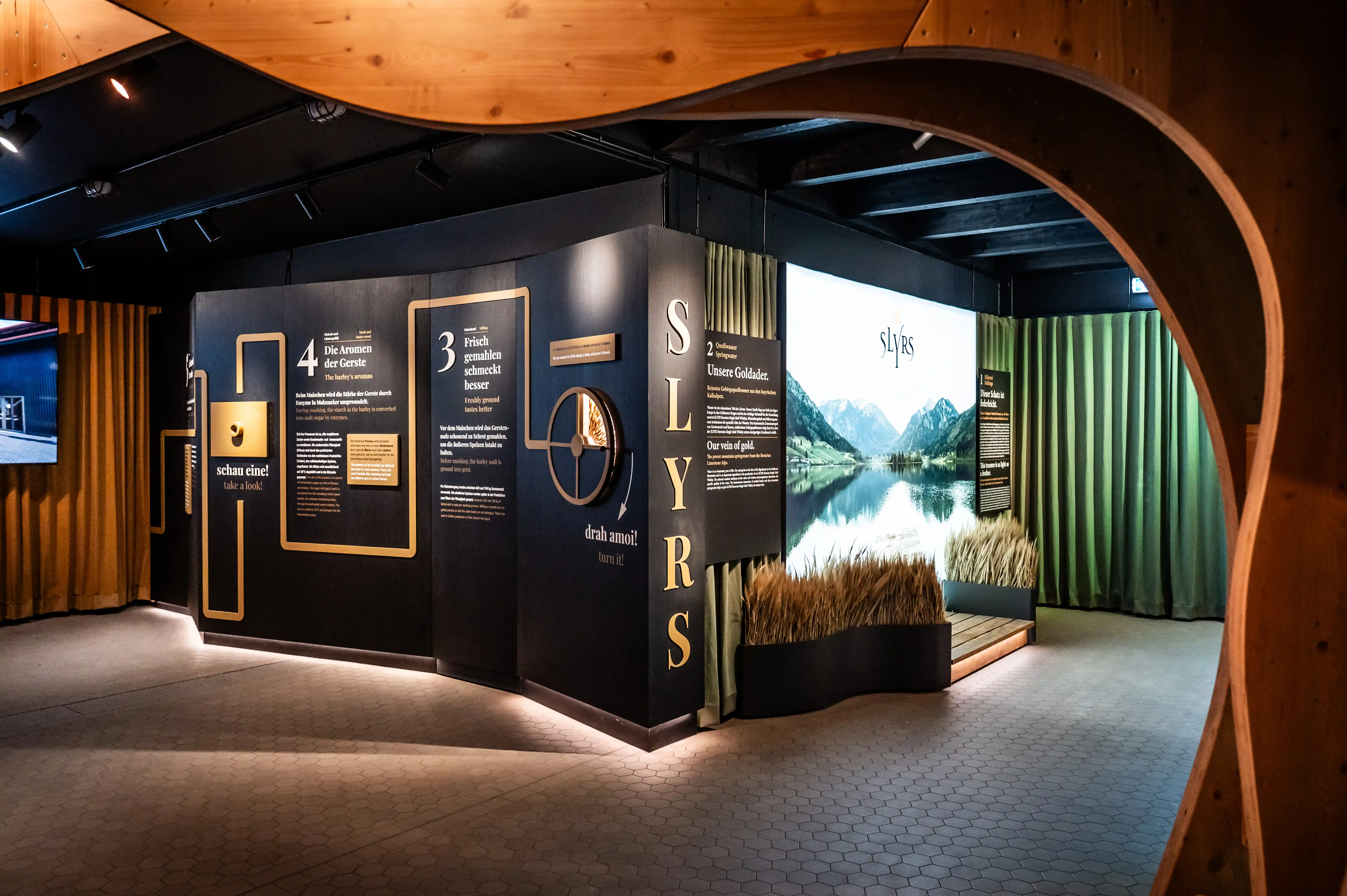 Dark exhibition walls with golden frames and numbered stations beside the glowing SLYRS lake panorama and green curtains.