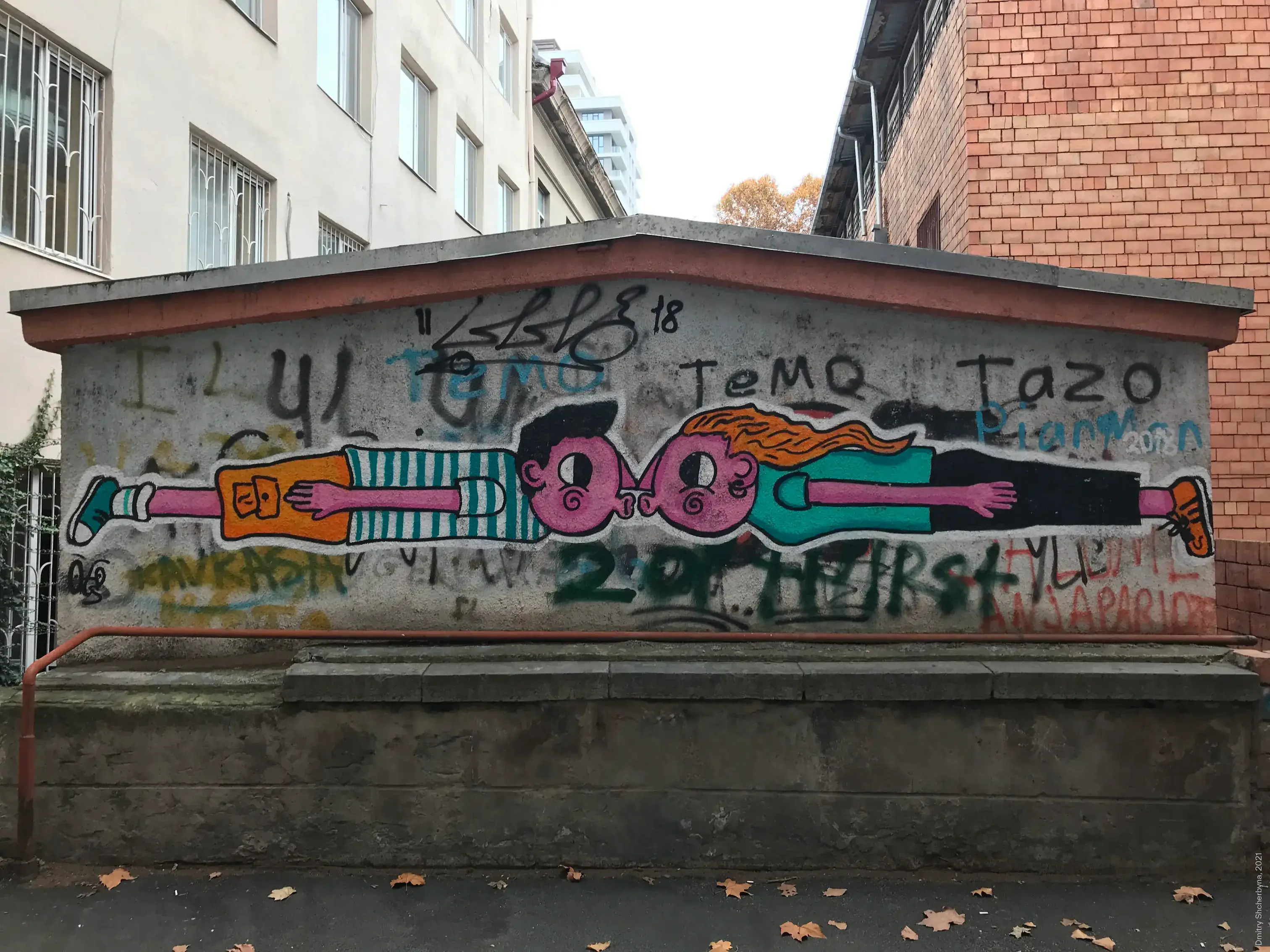 A simple, playful mural on a low concrete wall features two cartoon children, floating horizontally and kissing nose to nose. One has dark hair and is wearing an orange and striped outfit. The other has orange hair and wears teal and black clothing.