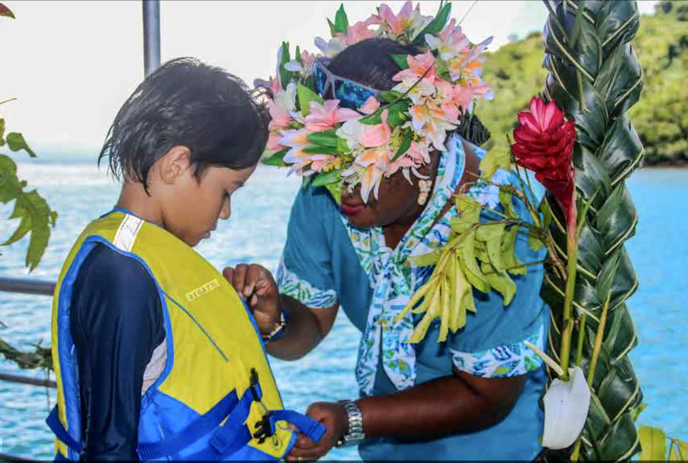 A Fijian lady staff member  with a flower crown helping a child secure his life jacket ready for a boat adventure
