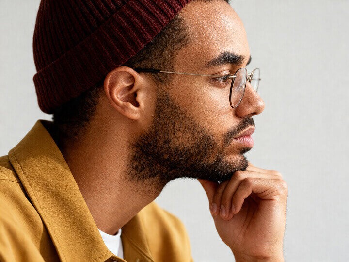 Smiling young designer wearing glasses and a burgundy beanie, dressed in a white t-shirt and caramel jacket, facing forward against a neutral background.