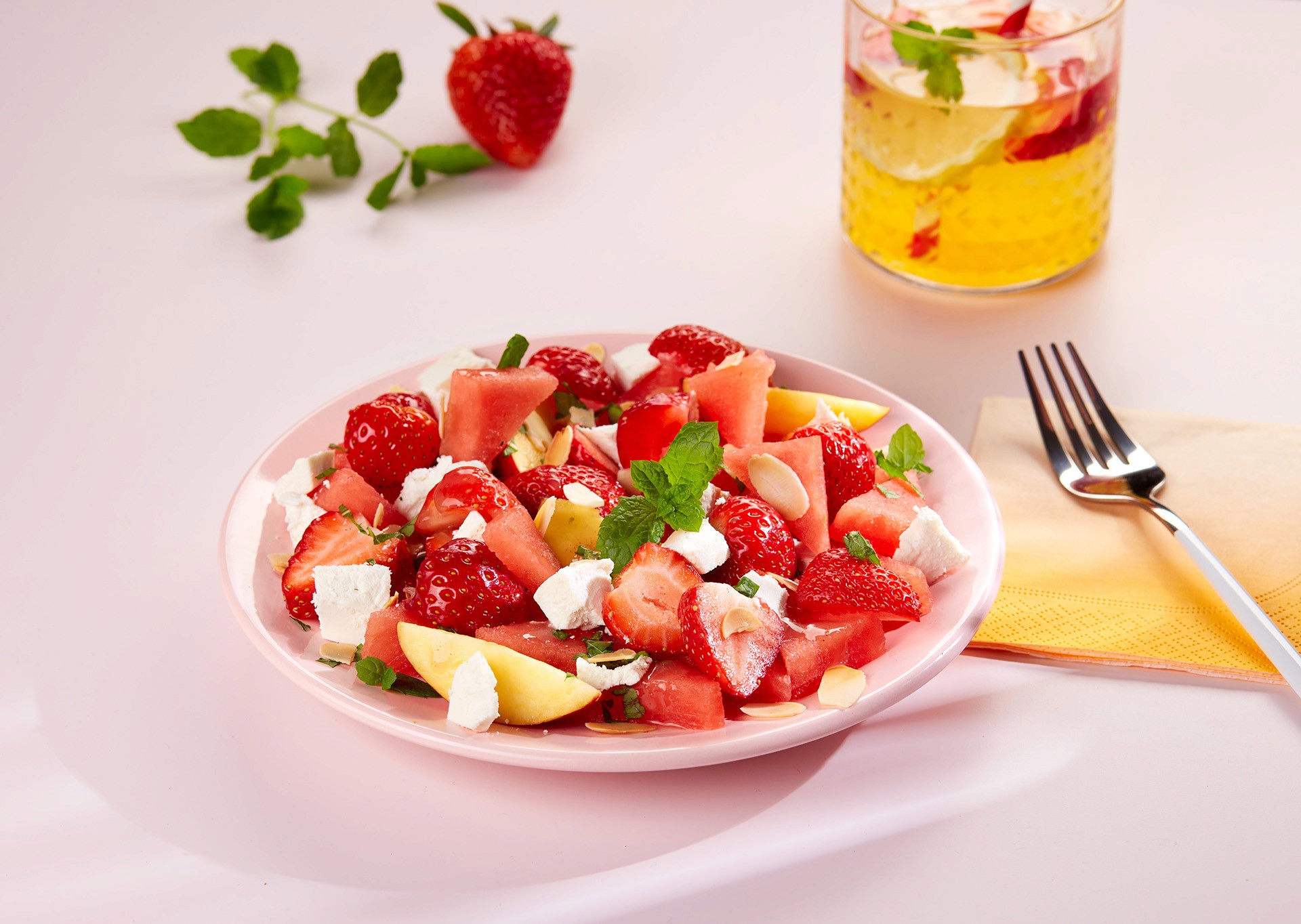 A vibrant fruit salad with strawberries, watermelon, feta cheese, and mint leaves is artfully arranged on a pink plate, accompanied by a fork, napkin, and a refreshing drink garnished with lemon and mint on a light pink table.