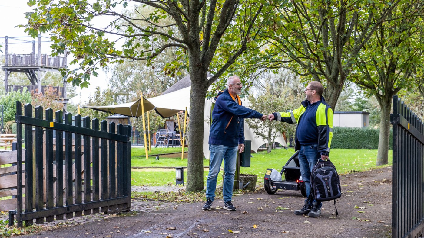 Two people engage in conversation near a stone gate, surrounded by trees and greenery on a pleasant day.