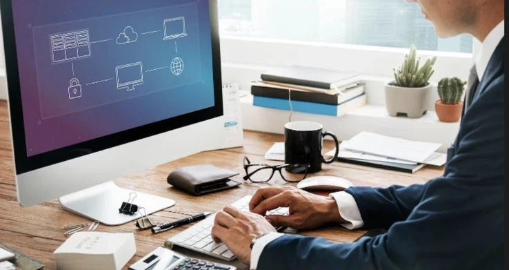 Mãos digitando num teclado, com um monitor exibindo ícones de documentos sobre a mesa