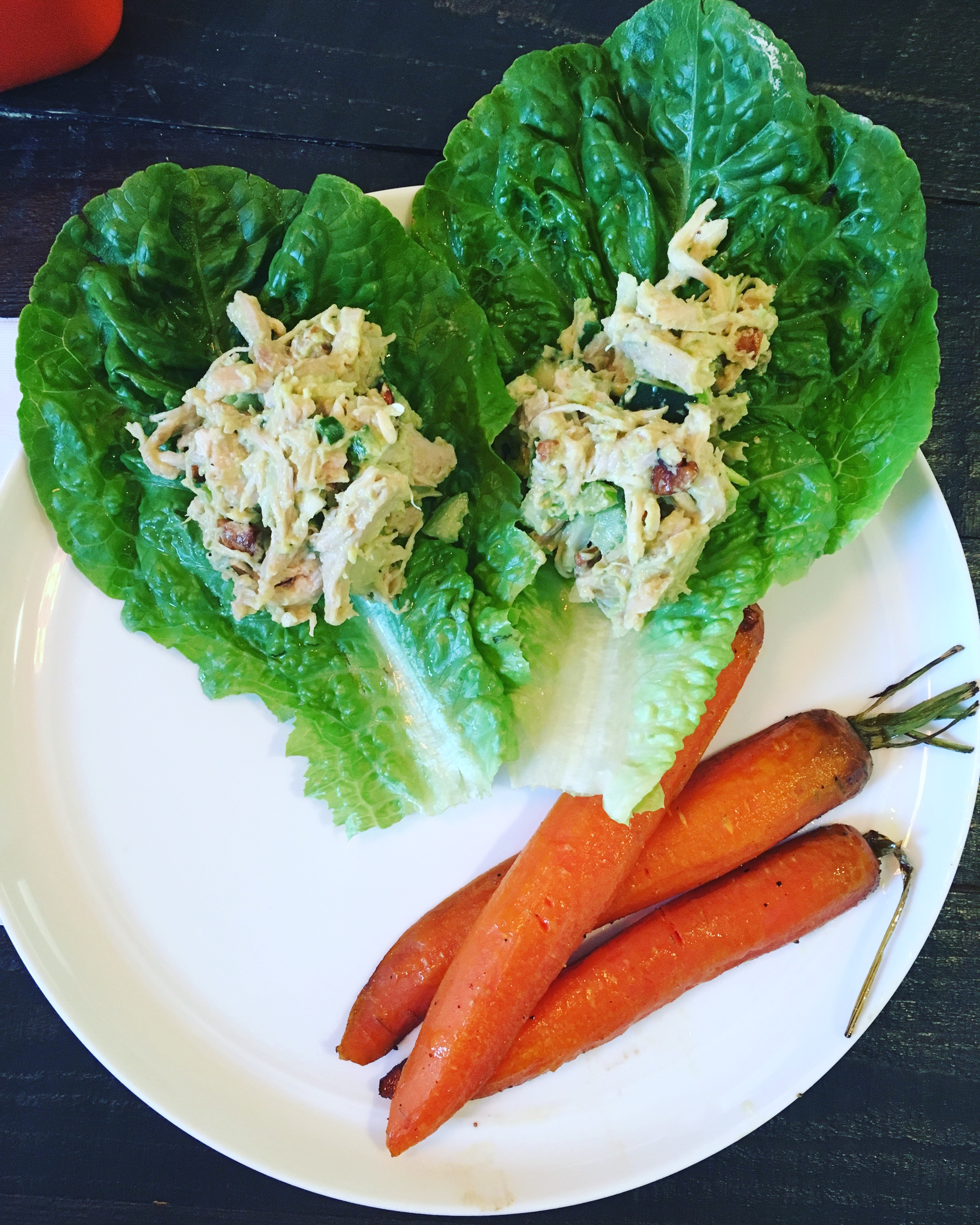 A white plate displays a fresh and healthy meal with two leafy green lettuce wraps filled with shredded chicken salad, accompanied by three roasted carrots.
