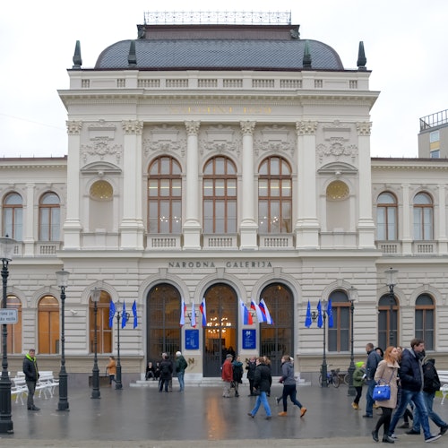 Хора, които се разхождат пред величествена сграда с арковидни прозорци, колони и множество знамена. На табелата пише "НАРОДНА ГАЛЕРИЯ."