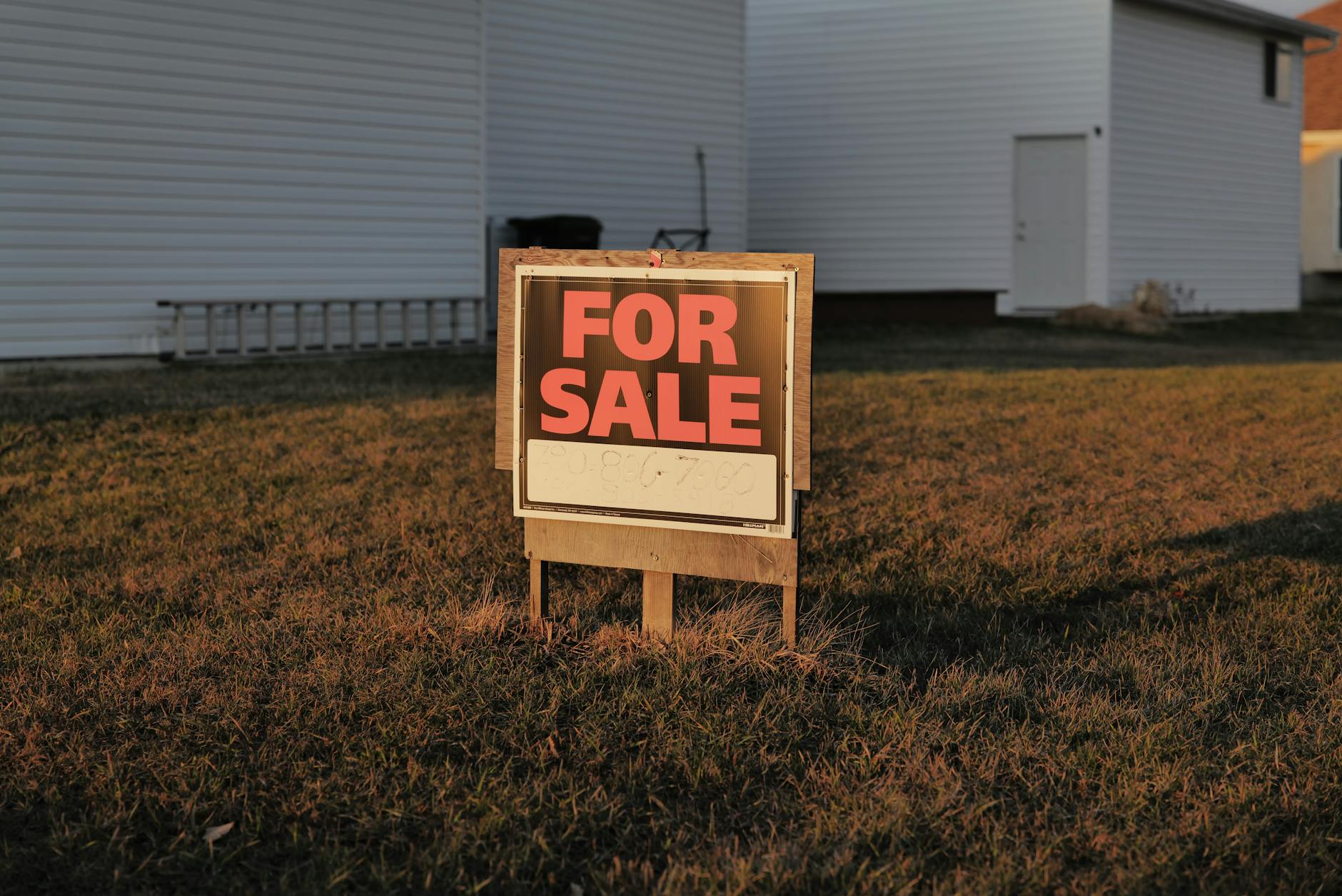 Wooden For Sale sign in a dry grass lawn outside a white residential house in warm afternoon light
