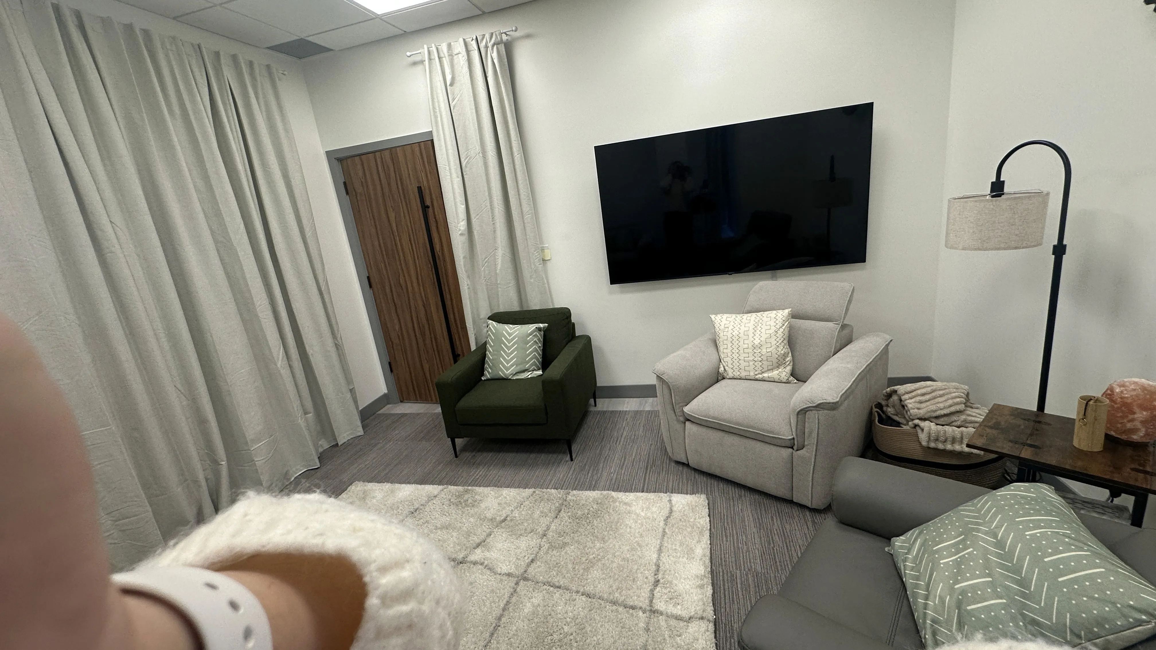 Lounge room with green, cream, and gray armchairs, a wall-mounted TV, and curtains