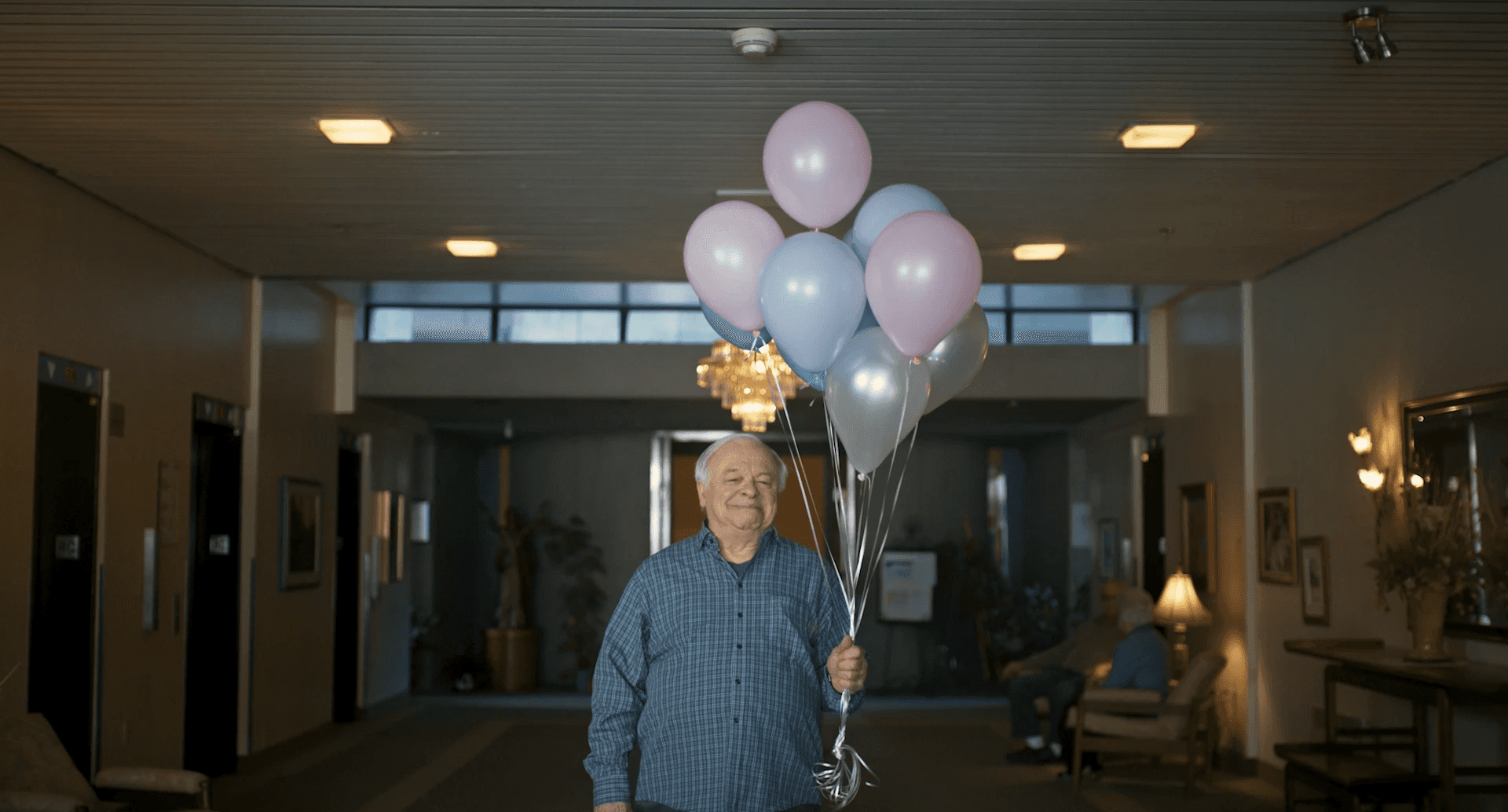Un homme agé se tient dans le hall d'entrée d'une maison de retraite, il tient un bouquet de balons couleurs argent et pastel. D'autres personnes agées sont assis et conversent en arrière plan.