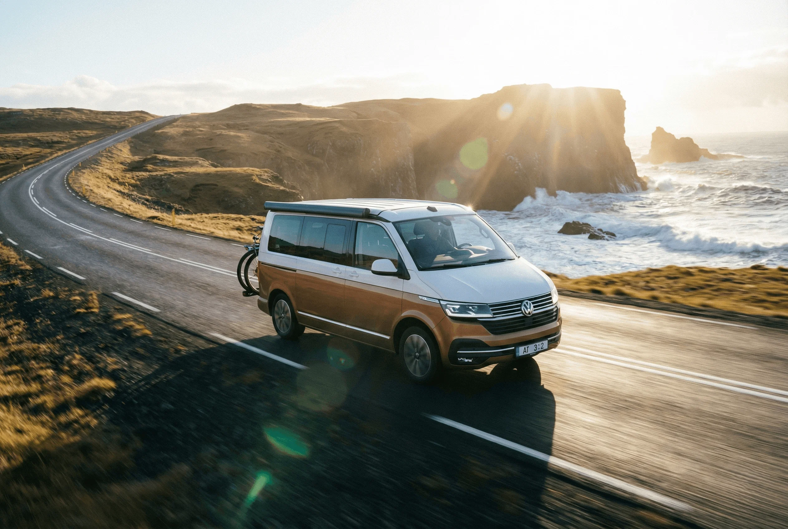 An orange camper van with its roof popped up parked on a grassy field in front of a large waterfall.