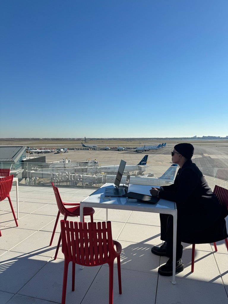 Ashleigh Warren works on a laptop at an outdoor table with red chairs, set against a clear blue sky and air strip runway landscape.