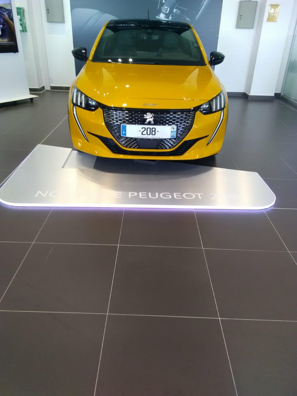 Une Peugeot 208 jaune exposée dans un showroom. Le design élégant et moderne de la voiture est mis en valeur par sa surface brillante et sa calandre stylée, qui lui confèrent une touche de sophistication.