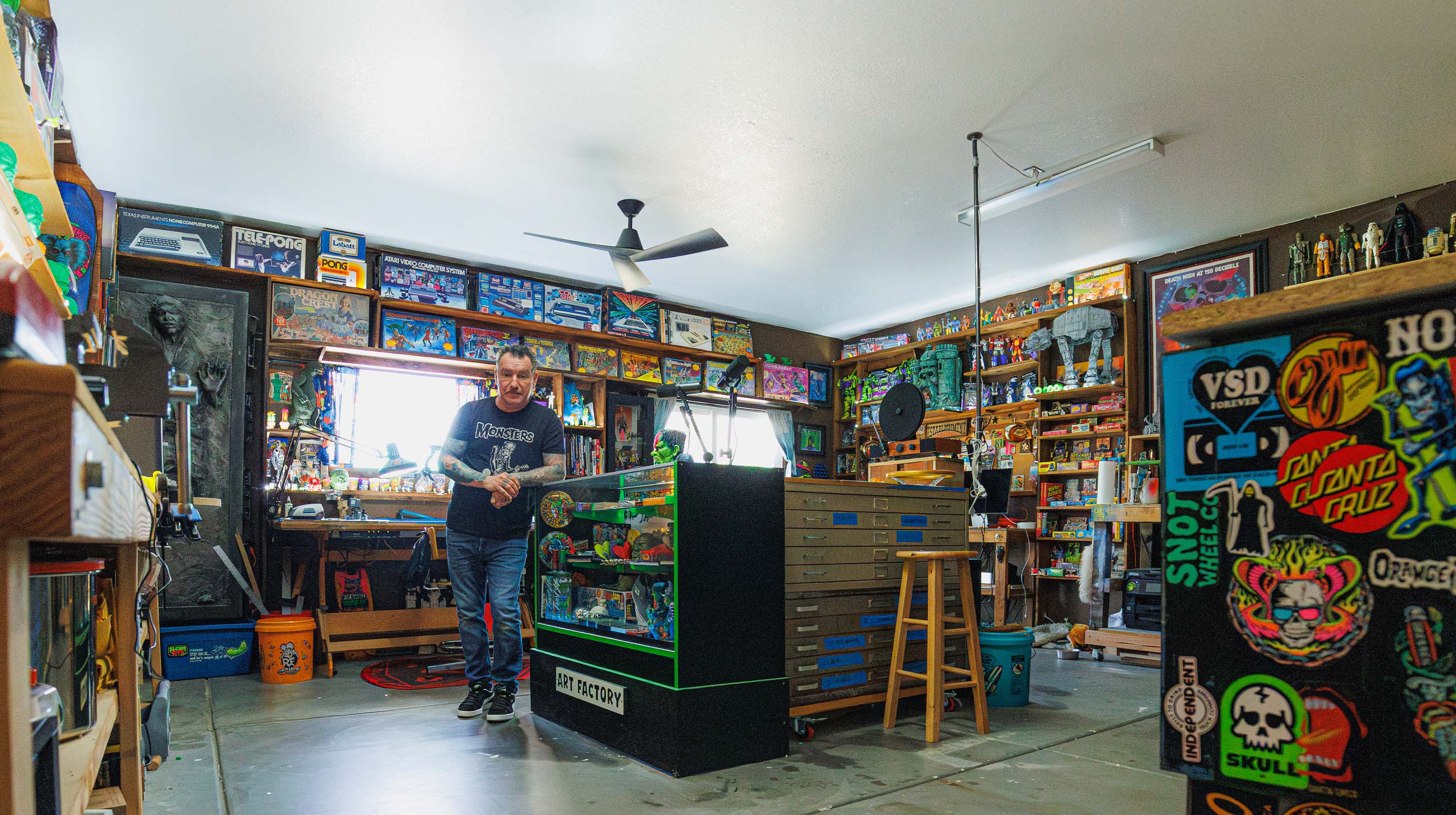 Artist Dirty Donny Gillies standing in his brightly lit studio filled with vintage toys, art supplies, and colorful collectibles, surrounded by shelves packed with models, posters, and pinball machines.