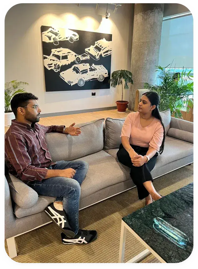A young man and woman engage in a serious conversation while seated on a light gray couch in a modern, well-lit office environment, featuring potted plants and abstract car artwork on the walls, representing a setting for Unbiased Counselling related to UG/PG Online Degree Programs with EazyGrad.