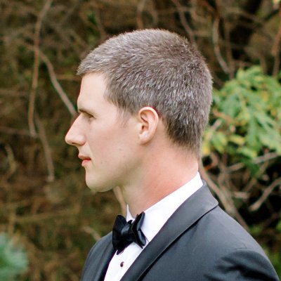 A man in a black tuxedo with a bow tie is standing outdoors against a blurred background of greenery and branches.