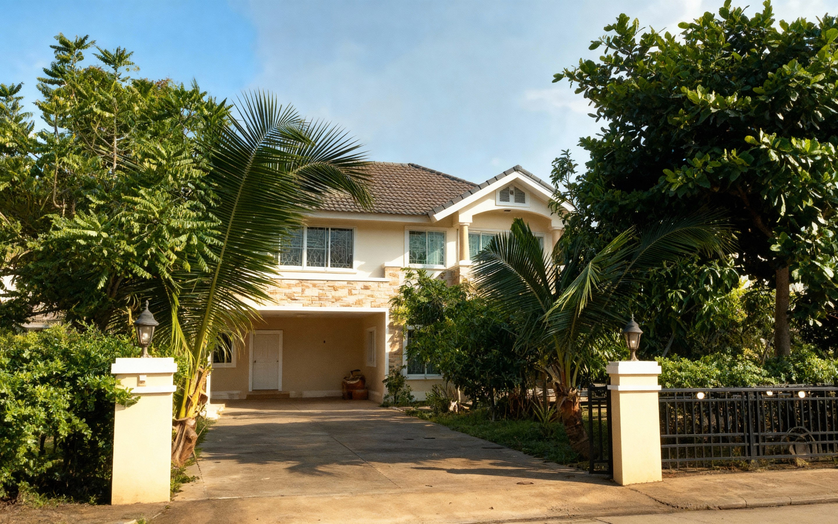 Two-story cream-colored house with a tiled roof surrounded by tropical trees and palms, set behind a gated driveway in the quiet, green suburb of Doi Saket, Chiang Mai.