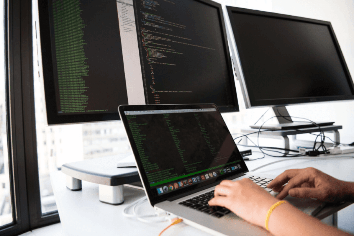 A developer typing on a laptop in front of two larger monitors on a white desk