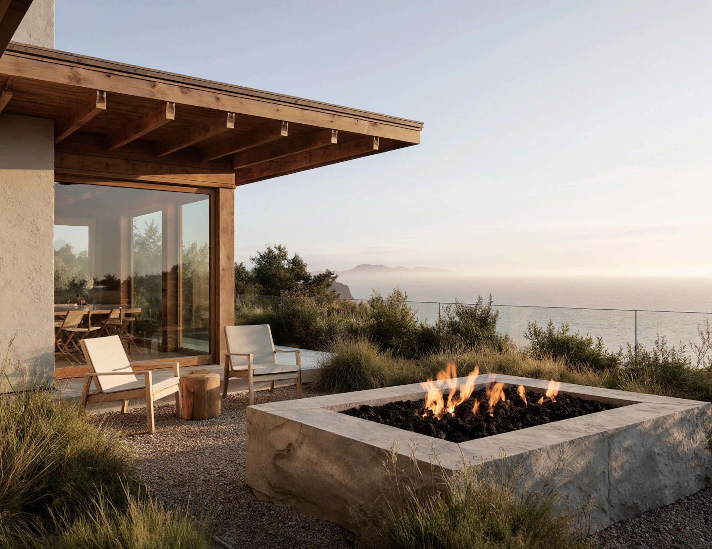 Contemporary coastal residence with an outdoor concrete fire pit, wooden lounge chairs, and native planting overlooking the ocean at dusk.