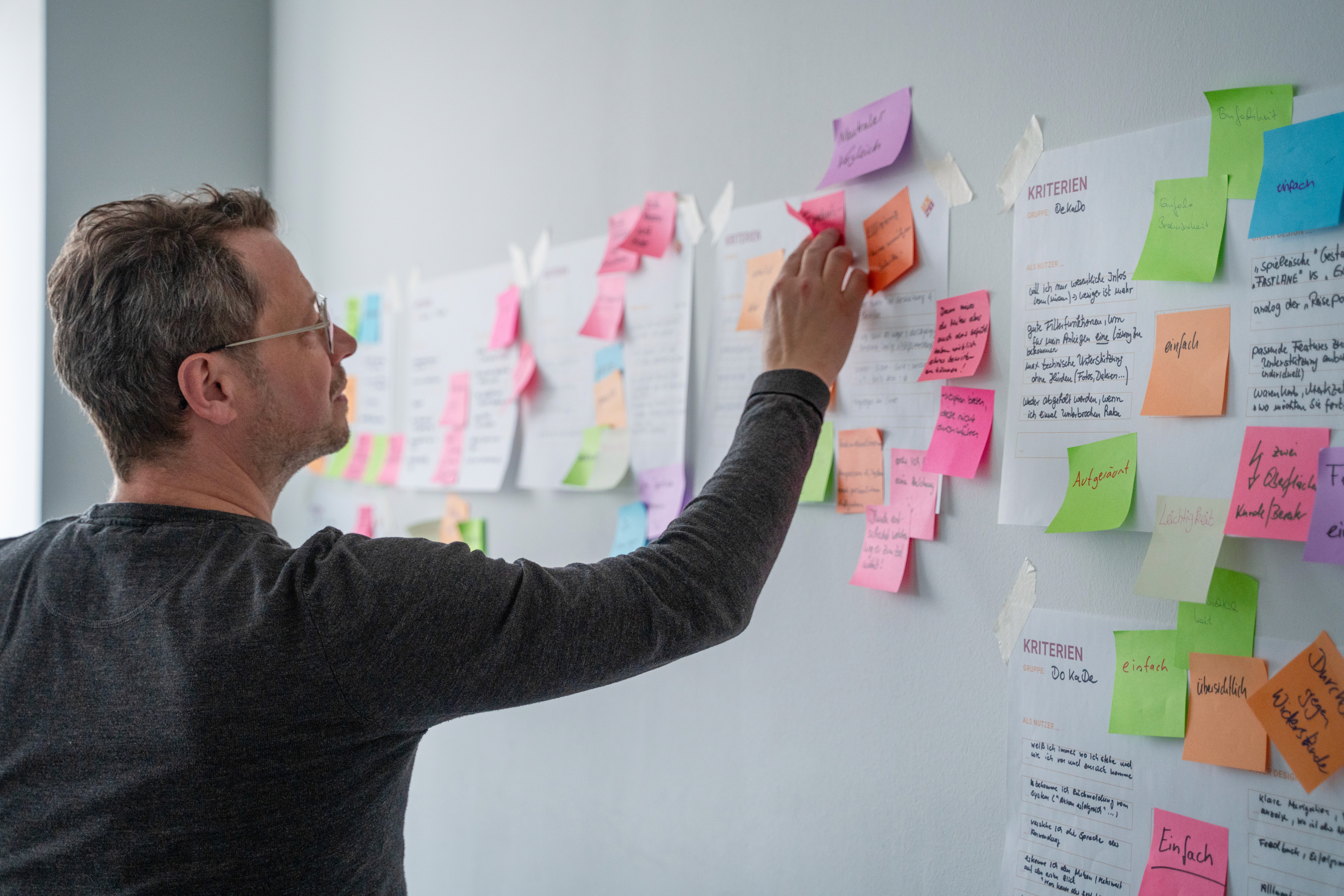 Person steht vor einer Wand mit zahlreichen farbigen Haftnotizen und Papieren und ordnet eine Haftnotiz an