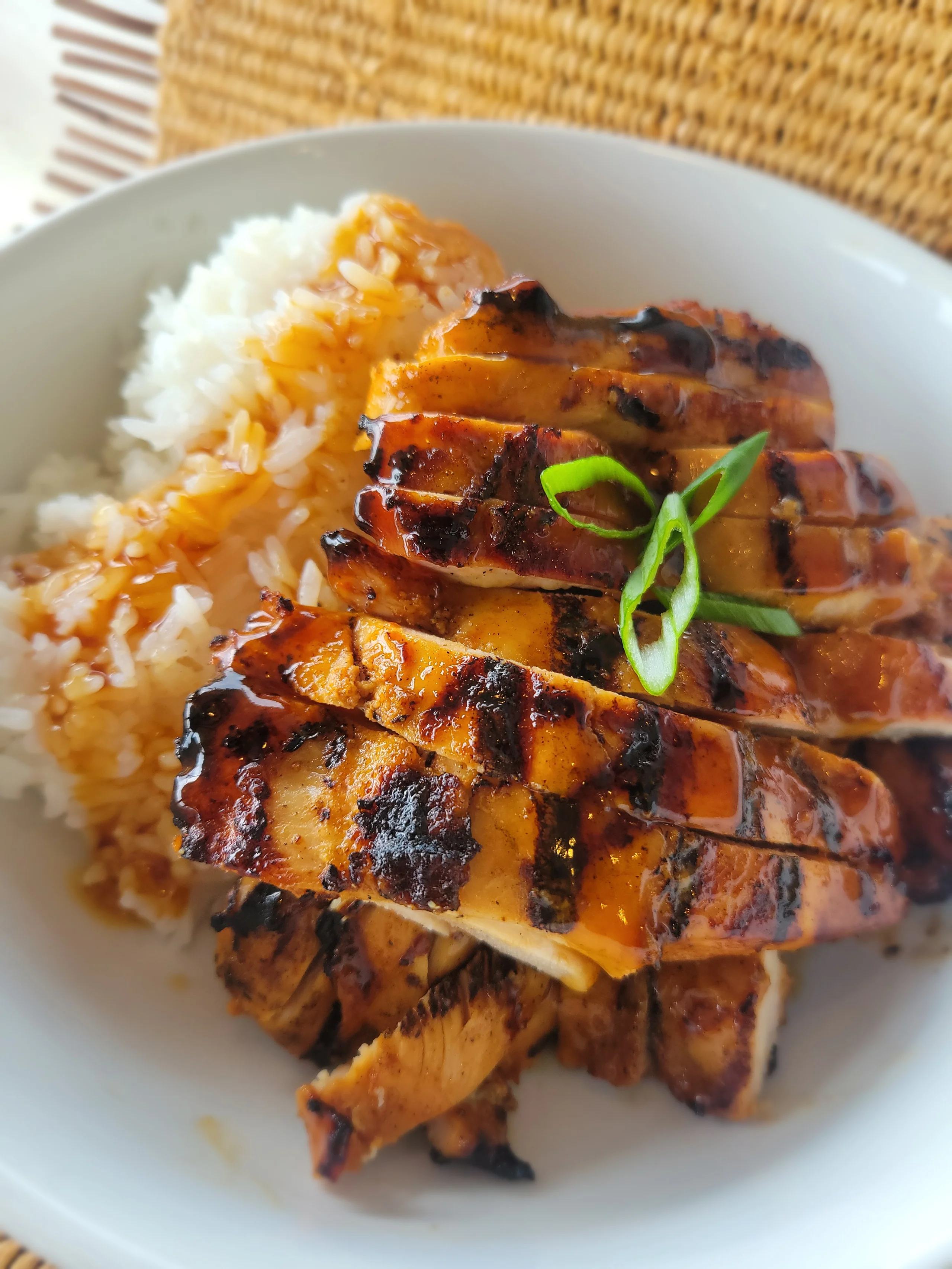 A white bowl filled with sliced, char-broiled Huli Huli chicken glazed in a dark sauce and garnished with green onions, served over a large portion of steamed white rice.